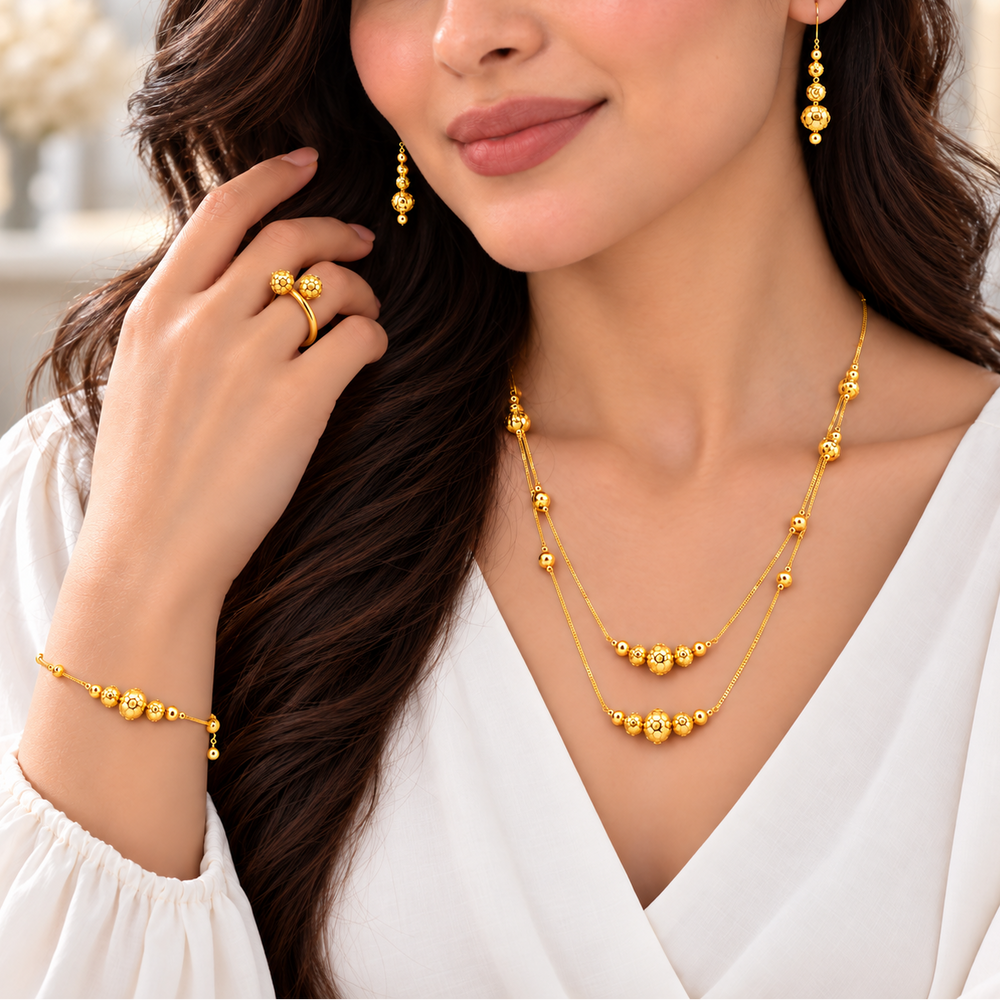 A woman wearing a gold jewelry set with a double-strand beaded necklace, matching drop earrings, and a bracelet, styled with a white blouse.